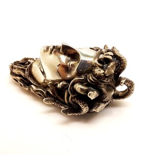 May include: A metallic pendant featuring a sculpted human face in profile with a smooth, polished finish. The hair is depicted as a cluster of textured, intertwined snakes with intricate scales, creating a high-contrast appearance between the smooth face and the detailed, dark, oxidized metal surroundings.