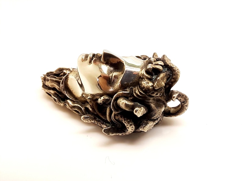 May include: A metallic pendant featuring a sculpted human face in profile with a smooth, polished finish. The hair is depicted as a cluster of textured, intertwined snakes with intricate scales, creating a high-contrast appearance between the smooth face and the detailed, dark, oxidized metal surroundings.