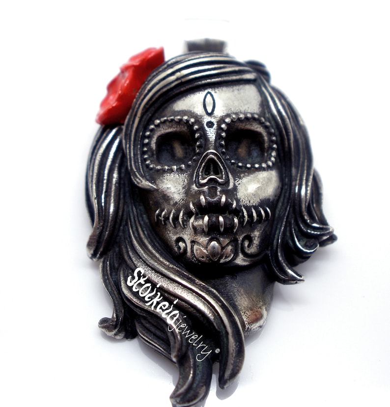 May include: A silver-toned pendant in the shape of a stylized skull with long hair and a red flower. The skull has intricate detailing around the eyes and mouth, and the text "Stoxie jewelry" is visible on the side.