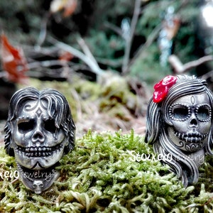 May include: Two silver-toned skull rings with intricate detailing. One ring features a male skull design, the other a female skull with a red bow. The rings are set against a backdrop of green moss and foliage, showcasing a Day of the Dead theme.