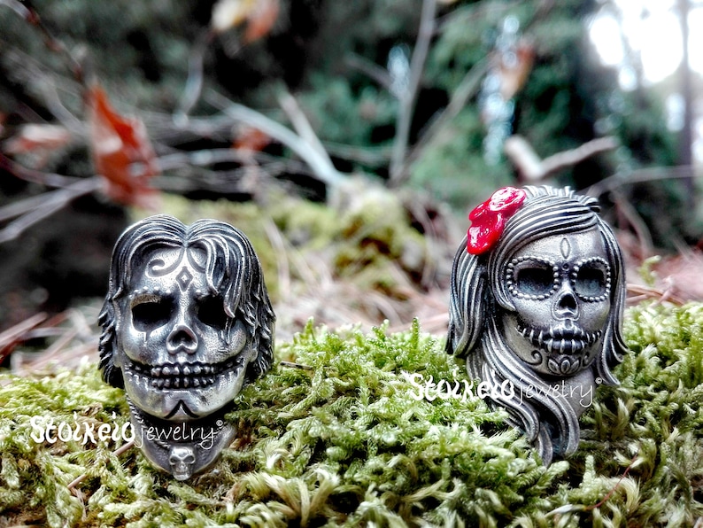 May include: Two silver-toned skull rings with intricate detailing. One ring features a male skull design, the other a female skull with a red bow. The rings are set against a backdrop of green moss and foliage, showcasing a Day of the Dead theme.