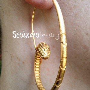 May include: A gold hoop earring featuring a textured surface with a repeating pattern of small indentations and a central decorative element shaped like a stylized head with beaded details. The text Stoixeio jewelry is visible on the earring.
