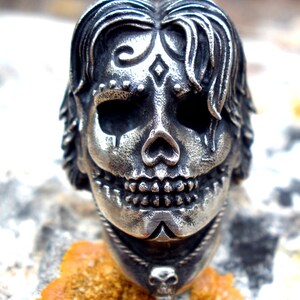 May include: A silver-toned skull ring with intricate details. The skull features stylized hair, decorative markings, and a smiling expression. A small skull pendant hangs below the main skull. The ring is set against a textured, orange-brown surface.