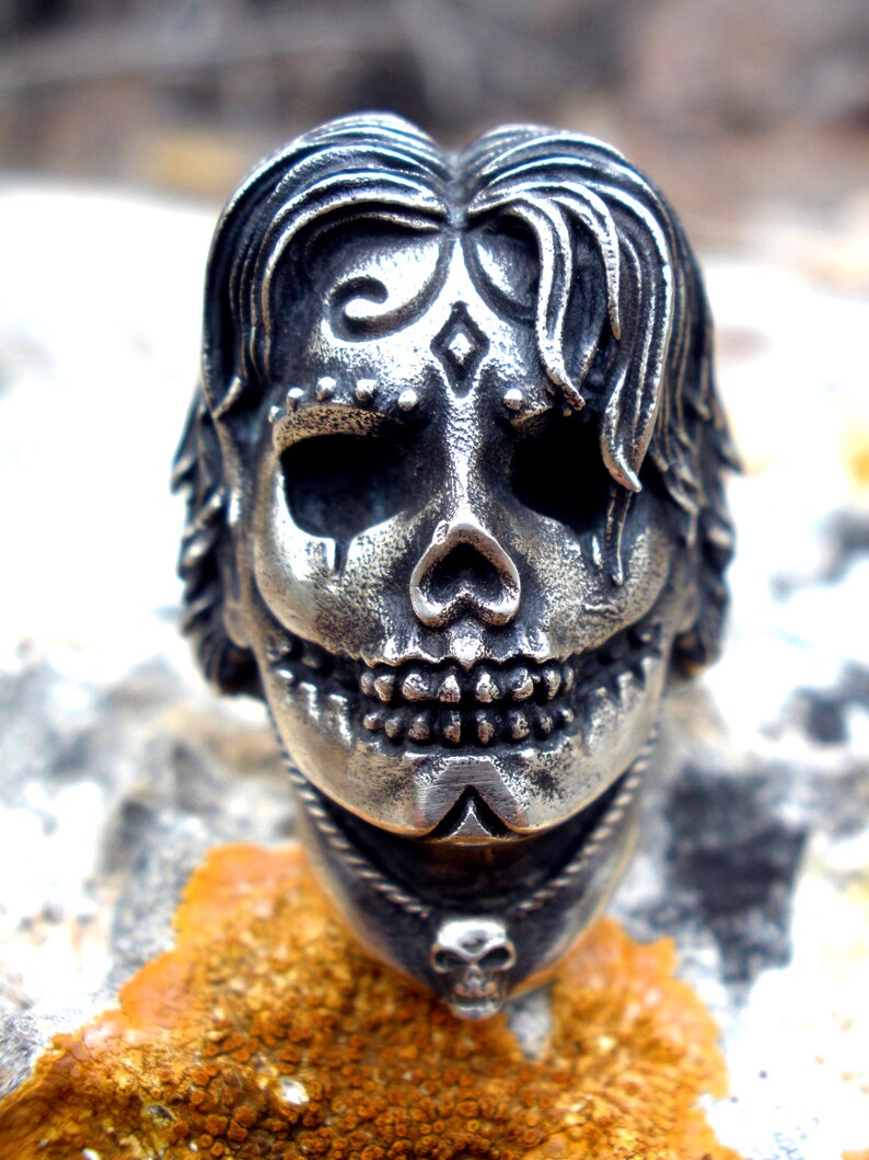 May include: A silver-toned skull ring with intricate details. The skull features stylized hair, decorative markings, and a smiling expression. A small skull pendant hangs below the main skull. The ring is set against a textured, orange-brown surface.