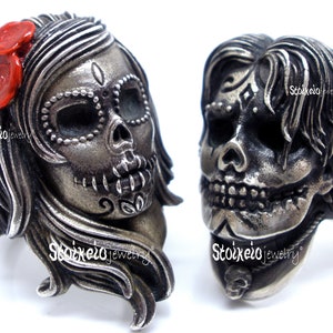 May include: Two silver-toned skull rings. One ring features a female skull with a red flower accent, the other a male skull. Both rings have intricate details and are designed in a Day of the Dead theme.