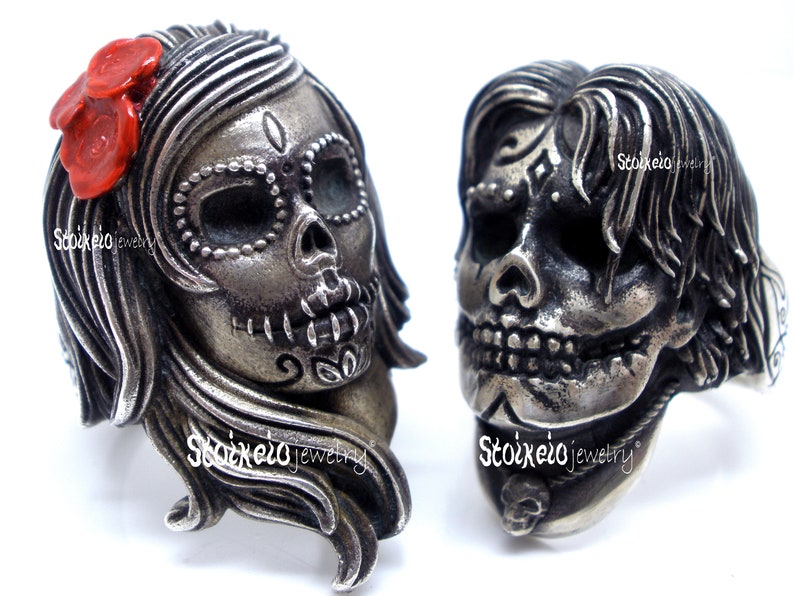May include: Two silver-toned skull rings. One ring features a female skull with a red flower accent, the other a male skull. Both rings have intricate details and are designed in a Day of the Dead theme.