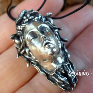 May include: A silver-toned pendant featuring a sculpted face with closed eyes and flowing, textured hair resembling snakes, suspended from a thin black cord. The text Stoixeio jewelry is visible in the lower right corner of the pendant.