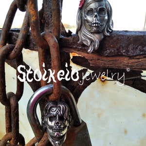 May include: Two silver Day of the Dead skull pendants, one on a padlock and the other resting on a rusty metal surface. The skull pendants have intricate details and are adorned with a red flower. The image includes the text "Stoikero jewelry".