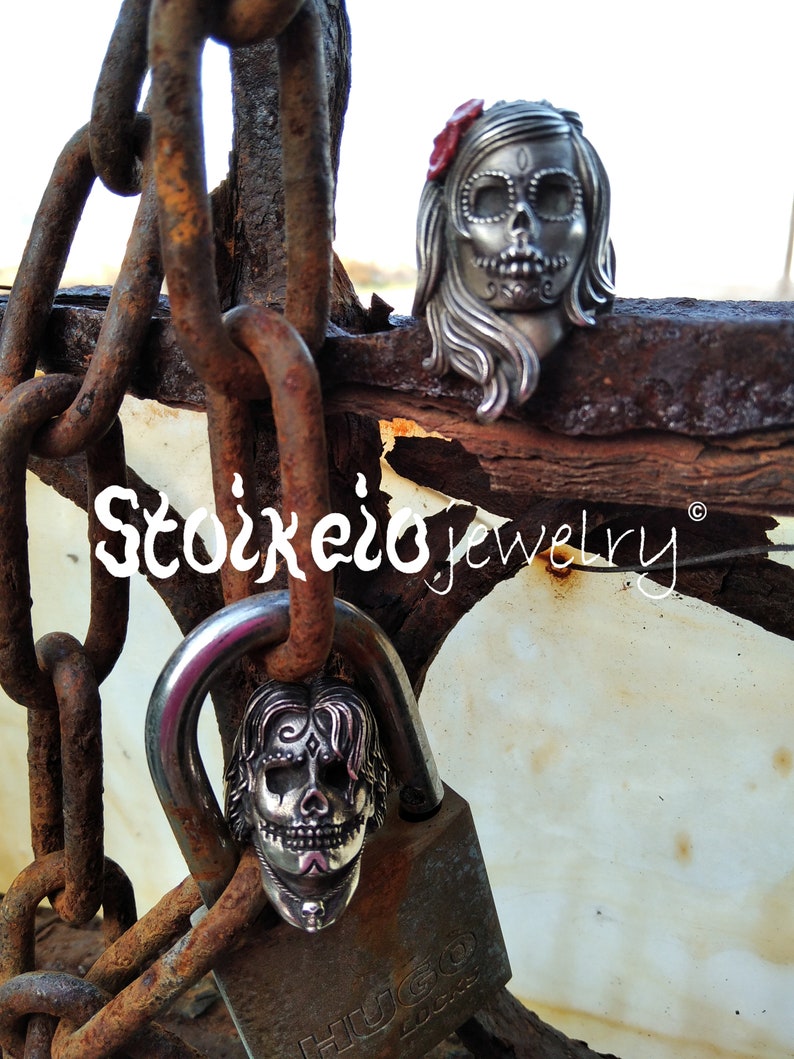 May include: Two silver Day of the Dead skull pendants, one on a padlock and the other resting on a rusty metal surface. The skull pendants have intricate details and are adorned with a red flower. The image includes the text "Stoikero jewelry".
