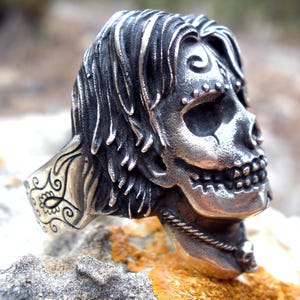 May include: A detailed silver skull ring with flowing hair and intricate facial features. The ring is set on a textured stone surface, showcasing the detailed craftsmanship of the jewelry. The skull design includes a decorative element on the ring's band.