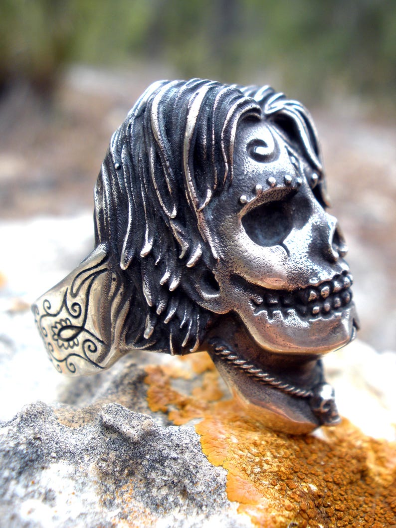 May include: A detailed silver skull ring with flowing hair and intricate facial features. The ring is set on a textured stone surface, showcasing the detailed craftsmanship of the jewelry. The skull design includes a decorative element on the ring's band.