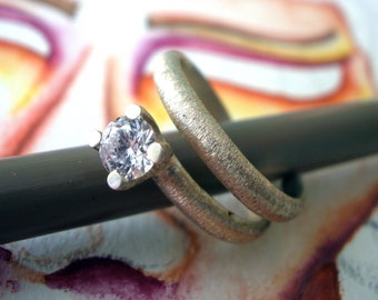 May include: A silver ring with a textured finish and a single, clear, round diamond. The ring is wrapped around a gray object.