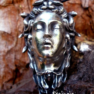 May include: A silver-toned pendant featuring a sculpted human face with closed eyes and parted lips, surrounded by snake-like forms in the hair. The piece is suspended from a black cord and includes the text Stoixeio jewelry at the bottom.