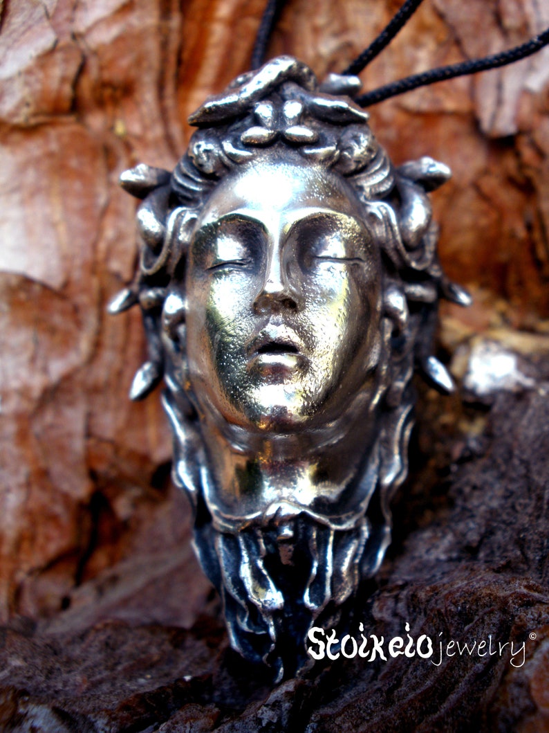 May include: A silver-toned pendant featuring a sculpted human face with closed eyes and parted lips, surrounded by snake-like forms in the hair. The piece is suspended from a black cord and includes the text Stoixeio jewelry at the bottom.