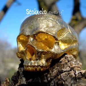 May include: A gold-colored ring shaped like a human skull with a textured, metallic finish. The text Stoixeio jewelry is visible above the skull. The skull features hollow eye sockets, a nasal cavity, and detailed teeth in a realistic anatomical form.