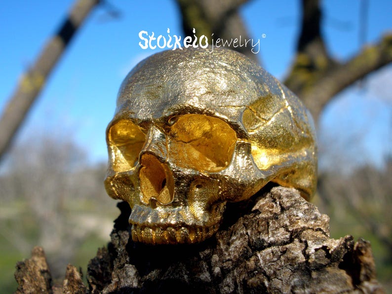 May include: A gold-colored ring shaped like a human skull with a textured, metallic finish. The text Stoixeio jewelry is visible above the skull. The skull features hollow eye sockets, a nasal cavity, and detailed teeth in a realistic anatomical form.