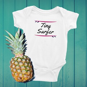 Puede incluir: Body de bebé blanco con el texto "Tiny Surfer" en escritura negra, enmarcado por un diseño de tabla de surf rosa y morado. Una piña madura con hojas verdes y una superficie texturizada está a la izquierda.