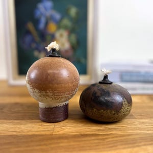 Può includere: Due lampade a olio in ceramica. Una è una lampada rotonda più grande con una glassa marrone e beige e una base scura. L'altra è una lampada rotonda più piccola con una glassa marrone scuro e verde. Entrambe hanno un supporto per stoppino nero e uno stoppino bianco.