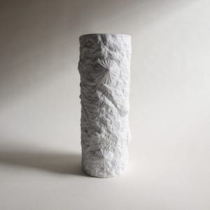 Puede incluir: Un jarrón blanco cilíndrico con una superficie texturizada y rugosa. El jarrón presenta una serie de diseños en relieve en forma de abanico. El diseño es minimalista y contemporáneo, adecuado para la decoración del hogar.