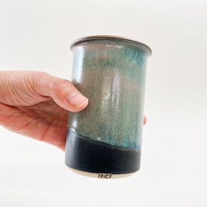 Könnte beinhalten: Eine Hand hält eine Keramikvase mit einer blauen und grünen Glasur. Die Vase hat einen schwarzen Streifen am Boden und einen weißen Sockel mit einer Markierung.