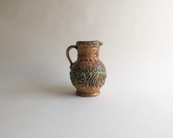 May include: A ceramic pitcher featuring a rounded body and a curved handle. The surface displays a textured, embossed pattern with a mix of earthy brown and muted green glazes. The neck has a scalloped design, while the lower section features intricate, swirling scrollwork motifs throughout the exterior.
