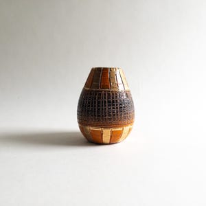 Op de afbeelding: Een kleine, ronde keramische vaas met een decoratieve buitenkant met afwisselende verticale strepen van licht- en donkerbruin aan de boven- en onderkant, gescheiden door een brede, getextureerde centrale band met een donkerbruin rasterpatroon.