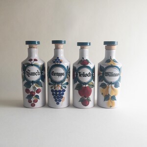 May include: Four white ceramic bottles with blue stoppers and labels. Each bottle features a different fruit illustration and text: "Kirsch", "Grappa", "Träsch", and "Williams". The bottles have a vintage aesthetic.