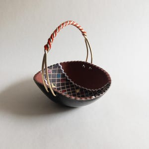 May include: A small, decorative basket with a black exterior and a brown interior. The rim features a patterned design with colorful squares. It has a twisted handle with red, white, and gold accents, and gold-colored metal supports.