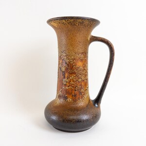 Könnte beinhalten: Eine braune und schwarze Keramikvase mit einer strukturierten Oberfläche und einem Henkel. Die Vase hat eine breite Öffnung und einen schmalen Hals.