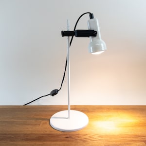 Puede incluir: Una lámpara de escritorio blanca con un brazo negro ajustable y una pantalla blanca. La lámpara está sobre una mesa de madera y está encendida, iluminando la mesa.