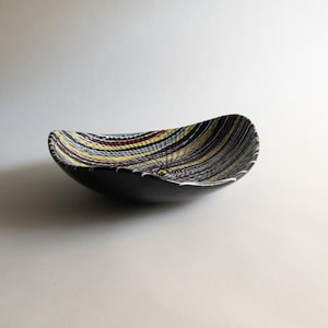 Op de afbeelding: Een zwarte keramische kom met een uniek, gestructureerd interieur. De binnenkant heeft een patroon van kleurrijke, horizontale strepen in geel, rood, blauw en wit. De kom heeft een gladde, matte zwarte buitenkant.