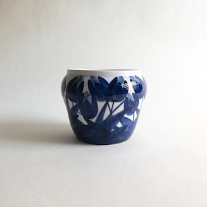 May include: A ceramic plant pot with a white rim and a dark blue floral design. The flowers are stylized tulip shapes. The pot is a rounded, squat shape, suitable for indoor plants. The background is plain white.