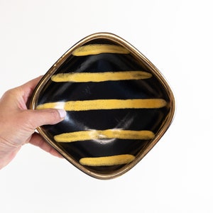 Puede incluir: Un plato cuadrado de cerámica con base negra y borde dorado. El plato tiene un patrón de rayas de líneas amarillas sobre la superficie negra.