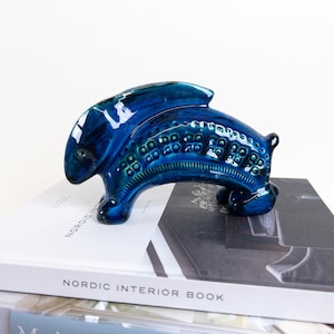 May include: A glossy, blue ceramic sculpture of a stylized animal, possibly a fish or a mythical creature. The sculpture has a textured surface with a dark teal hue and is displayed on a stack of books.