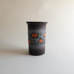 Puede incluir: Un jarrón cilíndrico de cerámica con una base marrón oscuro que se desvanece a gris con detalles en naranja y verde azulado. El jarrón tiene un borde ligeramente acampanado y presenta formas geométricas naranjas.