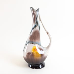 May include: A ceramic vase with a unique, abstract design in shades of brown, white, and blue. The vase has a wide, rounded base and a narrow neck with a curved handle.