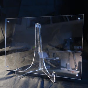 Puede incluir: Un expositor de acrílico transparente con una base rectangular y un soporte triangular. El expositor está diseñado para sostener un objeto plano, como una foto o una obra de arte.