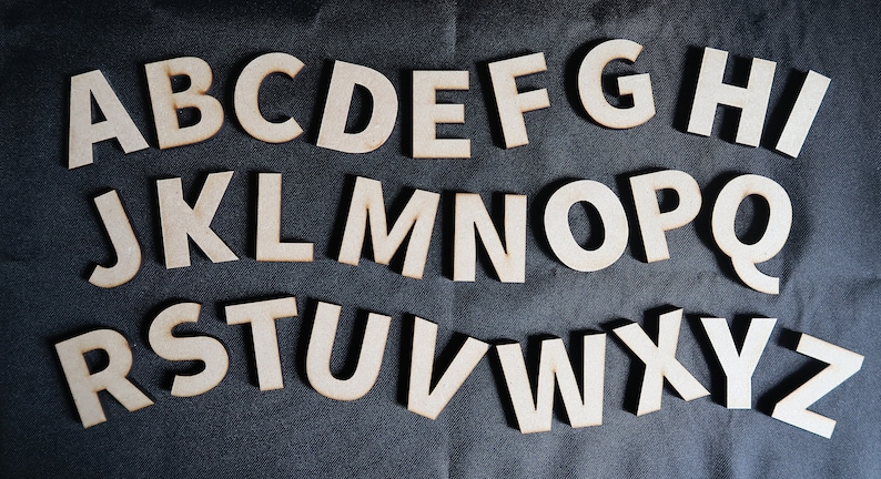 May include: Wooden letters of the alphabet, A through Z, in white, arranged in three rows on a dark gray fabric background.