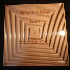 May include: A square wooden design with concentric square grooves. The text "Paul Williams Design" and "Square" are etched into the wood. The design includes measurements and the text "Please be aware" and "Due to the delicate nature of this product".
