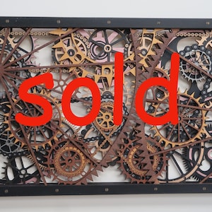 May include: Wooden wall art featuring a complex arrangement of gears and cogs in various sizes and colors, set within a black frame. The word "SOLD" is prominently displayed in red across the artwork.