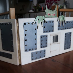 May include: A wooden, hinged box with metal plates and rivets. A green monster figure with red eyes and green hands is attached to the top. The box has a light wood finish and is designed with a fantasy theme.