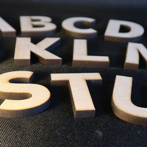 May include: Wooden letters of the alphabet, including A, B, C, D, E, F, J, K, L, M, N, R, S, T, U, and V. The letters are arranged in a random order and are on a dark surface.