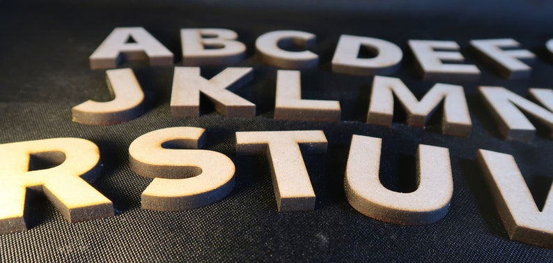 May include: Wooden letters of the alphabet, including A, B, C, D, E, F, J, K, L, M, N, R, S, T, U, and V. The letters are arranged in a random order and are on a dark surface.