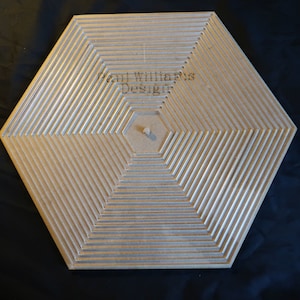 Op de afbeelding: Een zeshoekig houten bord met concentrische ringen die in het oppervlak zijn gegraveerd. Het bord heeft een centraal gat en de tekst "Paul Williams Designs" is bovenaan gegraveerd.