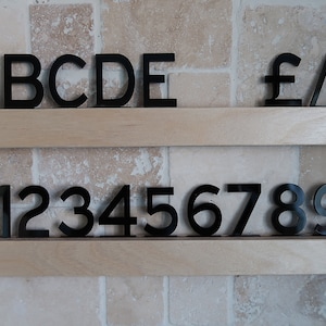 Puede incluir: Letras y números de plástico negro en dos estantes de madera sobre un fondo de azulejos beige. Las letras deletrean "ABCDE" y los números deletrean "0123456789". También hay símbolos para libra, barra oblicua y ampersand.
