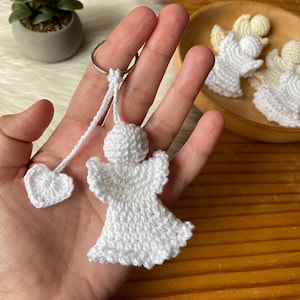 Puede incluir: Llavero de ángel blanco tejido a crochet con un pequeño dije de corazón blanco tejido a crochet unido al anillo. El llavero se sostiene en una mano.