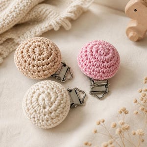 May include: Three crochet pacifier clips in beige, pink, and off-white. Each clip features a round, crocheted design attached to a metal clip. A wooden toy horse and a crocheted blanket are in the background.