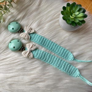 May include: Two mint green crocheted baby teethers with white crocheted bows and silver clips.