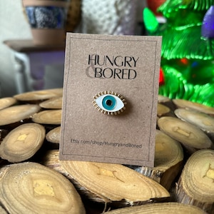 May include: A white and blue enamel pin with a gold outline, depicting a stylized eye. The pin is on a brown card with the text "HUNGRY & BORED" and "Etsy.com/shop/HungryandBored".