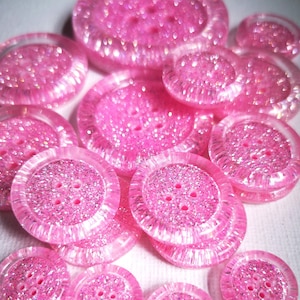 Könnte beinhalten: Eine Sammlung von rosa Glitzerknöpfen in verschiedenen Größen und Designs. Die Knöpfe haben eine glänzende, schillernde Oberfläche.